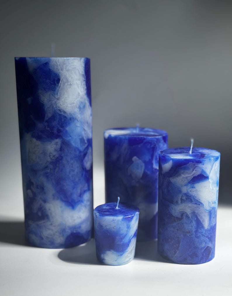 May include: Four blue and white marbled pillar candles of varying heights. The candles have a marbled effect with shades of blue and white. The wicks are visible on top. The candles are cylindrical and stand on a white surface.