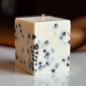 K&ouml;nnte beinhalten: Quadratische, cremefarbene Kerze mit dunkelbraunen Sprenkeln. Die Kerze hat einen einzelnen Docht und steht auf einer wei&szlig;en Oberfl&auml;che. Das Design der Kerze erinnert an einen K&auml;seblock.