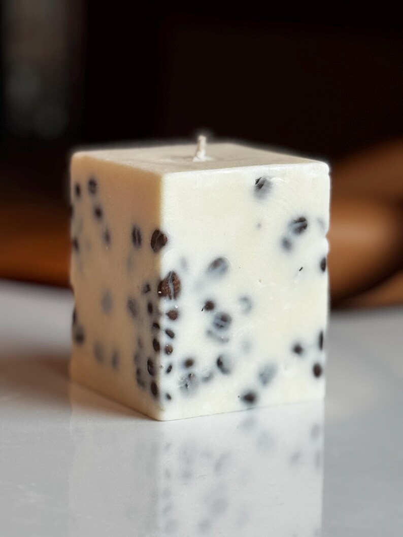 K&ouml;nnte beinhalten: Quadratische, cremefarbene Kerze mit dunkelbraunen Sprenkeln. Die Kerze hat einen einzelnen Docht und steht auf einer wei&szlig;en Oberfl&auml;che. Das Design der Kerze erinnert an einen K&auml;seblock.