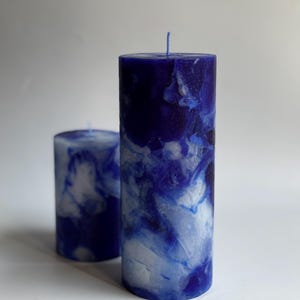 May include: Two pillar candles with a marbled blue and white design. The larger candle is approximately 6 inches tall, and the smaller one is about 3 inches tall. Both candles have a wick at the top and are cylindrical in shape. The background is plain white.
