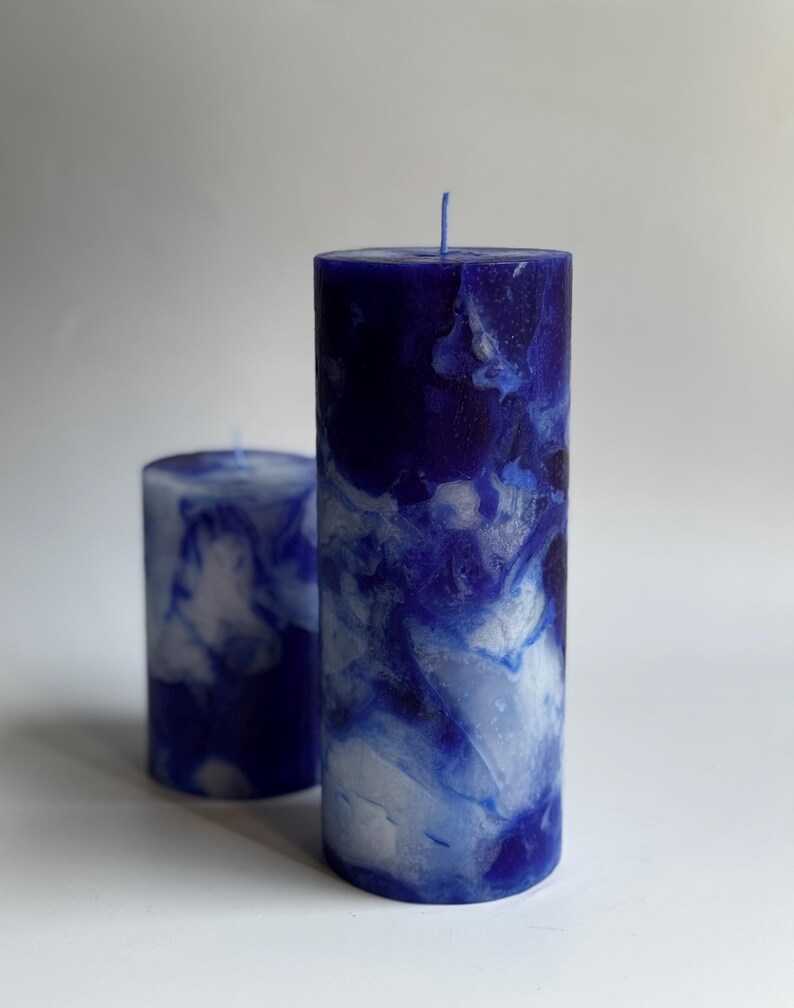 May include: Two pillar candles with a marbled blue and white design. The larger candle is approximately 6 inches tall, and the smaller one is about 3 inches tall. Both candles have a wick at the top and are cylindrical in shape. The background is plain white.