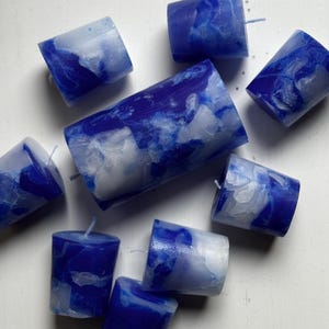 May include: A collection of blue and white marbled votive candles. The candles are cylindrical with a flat top and a wick. The color pattern resembles a cloudy sky. The candles are scattered on a white surface.