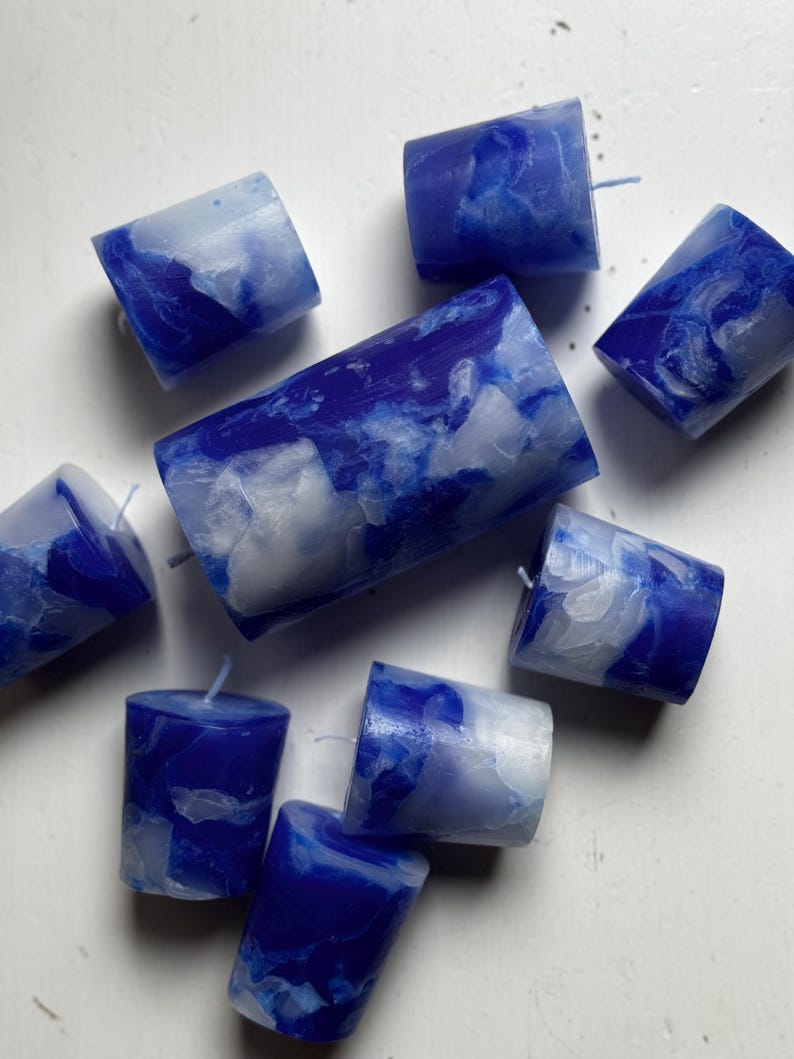 May include: A collection of blue and white marbled votive candles. The candles are cylindrical with a flat top and a wick. The color pattern resembles a cloudy sky. The candles are scattered on a white surface.