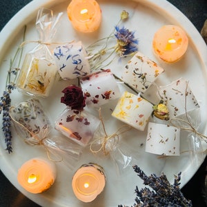 K&ouml;nnte beinhalten: Sortiment kleiner, runder, wei&szlig;er Kerzen mit getrockneten Blumen und Bl&uuml;tenbl&auml;ttern, die in das Wachs eingebettet sind. Die Kerzen sind auf einer wei&szlig;en Marmorfl&auml;che angeordnet.