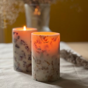May include: Two white pillar candles with embedded dried flowers and a warm glow. The candles are on a white linen tablecloth.