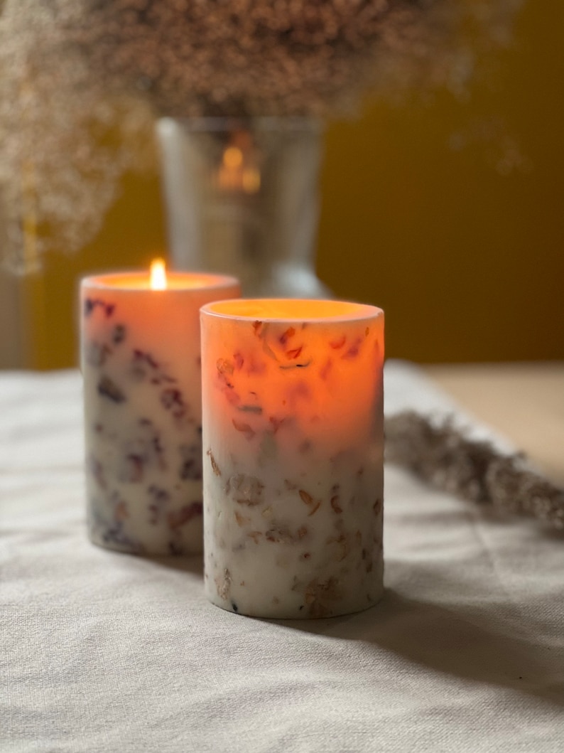 May include: Two white pillar candles with embedded dried flowers and a warm glow. The candles are on a white linen tablecloth.