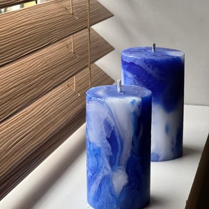 May include: Two pillar candles with a marbled blue and white design. The candles are cylindrical with a wick at the top. They are placed on a white surface near a wooden window blind.
