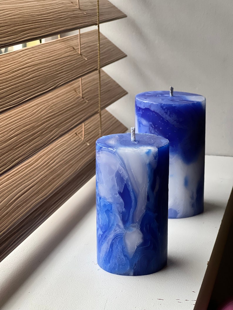 May include: Two pillar candles with a marbled blue and white design. The candles are cylindrical with a wick at the top. They are placed on a white surface near a wooden window blind.