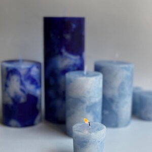 May include: A collection of blue and white marbled pillar candles of varying heights. One small candle is lit, with a visible flame. The candles have a marbled effect, creating a cloudy appearance. The color palette is primarily shades of blue and white.