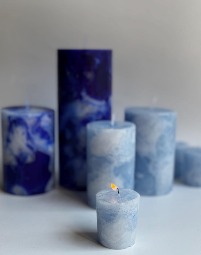 May include: A collection of blue and white marbled pillar candles of varying heights. One small candle is lit, with a visible flame. The candles have a marbled effect, creating a cloudy appearance. The color palette is primarily shades of blue and white.