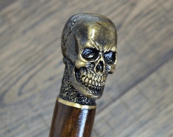 May include: A wooden walking stick with a detailed skull head made of gold-colored metal. The skull has a menacing expression with a wide open mouth and sharp teeth.