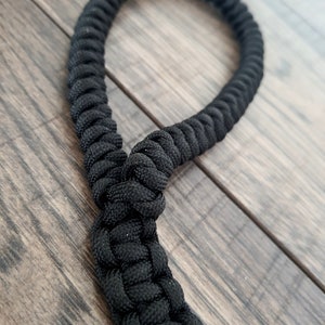Pu&ograve; includere: Un cinturino in paracord nero con un nodo al centro. Il cinturino &egrave; fatto di corda intrecciata e si trova su una superficie di legno.