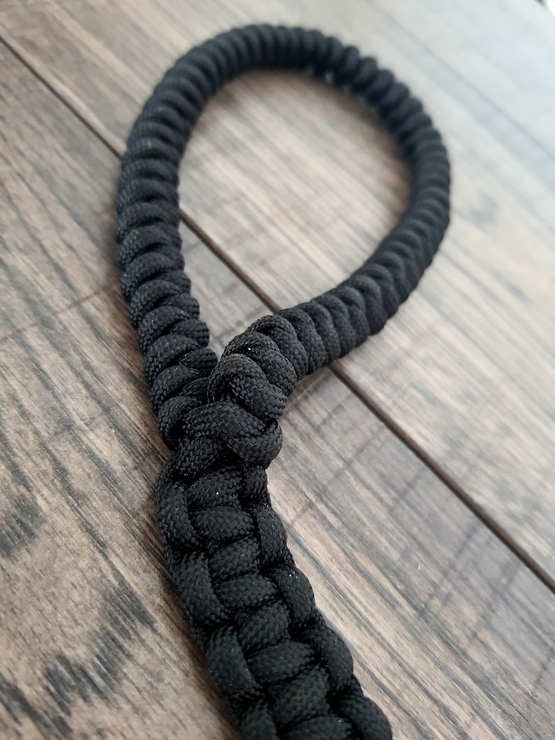Pu&ograve; includere: Un cinturino in paracord nero con un nodo al centro. Il cinturino &egrave; fatto di corda intrecciata e si trova su una superficie di legno.