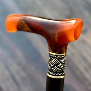 Op de afbeelding: Een wandelstokhandvat met een gepolijste, doorschijnende oranje en donkerbruine harsbovenkant. Onder het handvat bevindt zich een metalen kraag met een gedetailleerd, gevlochten touwpatroon in een zilverkleurige afwerking, verbonden met een gladde, donkere schacht.