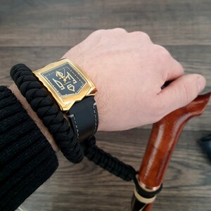 Pu&ograve; includere: Un orologio dorato con cinturino in pelle nera e quadrante nero con dettagli dorati. L'orologio &egrave; indossato su un polso con un braccialetto intrecciato nero.