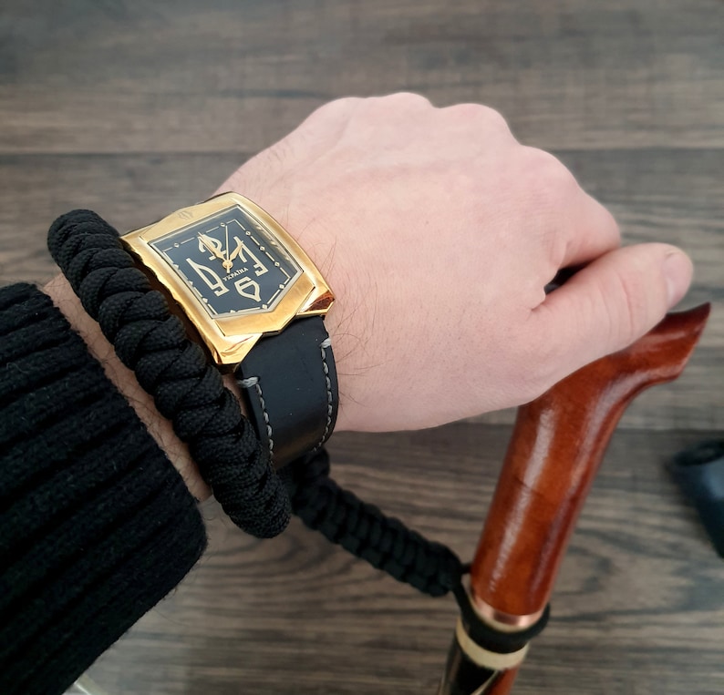Pu&ograve; includere: Un orologio dorato con cinturino in pelle nera e quadrante nero con dettagli dorati. L'orologio &egrave; indossato su un polso con un braccialetto intrecciato nero.