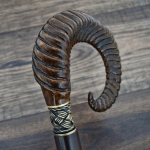 Peut inclure: Une canne de marche en bois avec une poignée sculptée en forme de corne de bélier. La poignée est de couleur brun foncé et présente un motif en spirale détaillé. La canne est décorée d'une bande dorée.