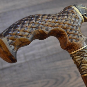 Puede incluir: Un bast&oacute;n de madera con un mango en forma de calavera y un dise&ntilde;o de cuerda trenzada. El mango est&aacute; tallado intrincadamente con un patr&oacute;n tejido y presenta una banda dorada.