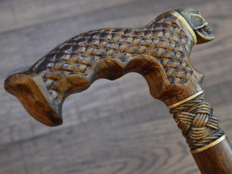 Puede incluir: Un bast&oacute;n de madera con un mango en forma de calavera y un dise&ntilde;o de cuerda trenzada. El mango est&aacute; tallado intrincadamente con un patr&oacute;n tejido y presenta una banda dorada.