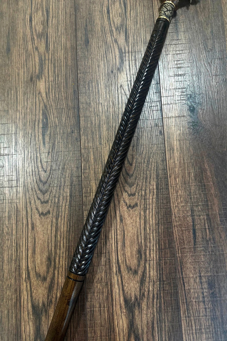 Puede incluir: Un bast&oacute;n de madera con un acabado marr&oacute;n oscuro y un dise&ntilde;o tallado. El bast&oacute;n tiene una parte superior de metal con un dise&ntilde;o decorativo.