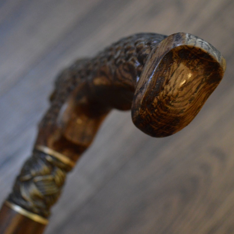 Puede incluir: Un bast&oacute;n de madera con un mango tallado y una banda met&aacute;lica decorativa. El mango es de color marr&oacute;n oscuro y tiene una veta de madera natural.