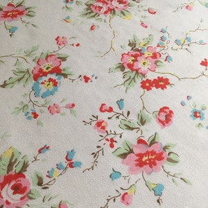 以下が含まれることがあります： 花柄の生地のクローズアップ。ピンクと赤のバラのクラスター、小さな青い花、緑の葉がクリーム色の背景にデザインされています。繊細な茶色のつるが花の要素をつないでいます。