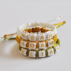 Peut inclure: Trois bracelets d'amitié avec les noms "Noah", "Adam" et "Elias" écrits en lettres dorées sur des perles blanches. Les bracelets sont faits de ficelle blanche, jaune et verte.