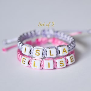 May include: Two handmade friendship bracelets. The top bracelet is lavender and white, spelling "ISLA". The bottom bracelet is pink and white, spelling "ELISE". Both bracelets have gold lettering. The text "Set of 2" is above the bracelets.