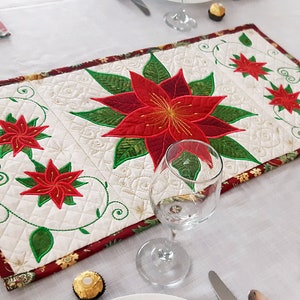 Op de afbeelding: Een witte tafelrunner met een rood en groen kerststerrenontwerp. De runner is afgewerkt met een rode rand en heeft een gestikt patroon.