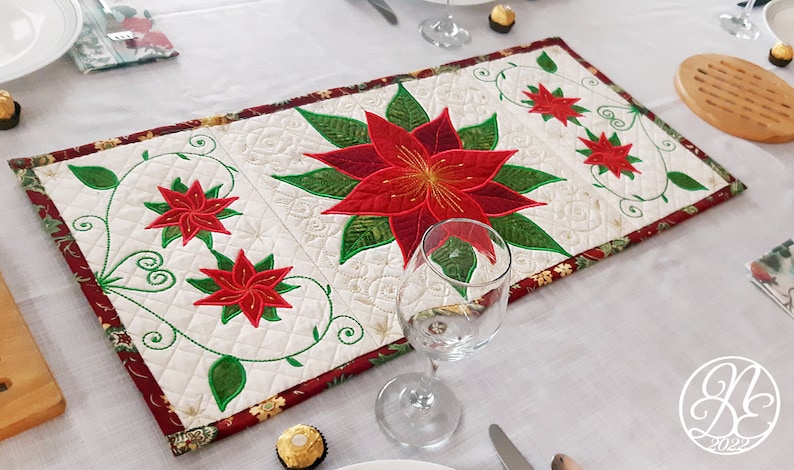 Op de afbeelding: Een witte tafelrunner met een rood en groen kerststerrenontwerp. De runner is afgewerkt met een rode rand en heeft een gestikt patroon.