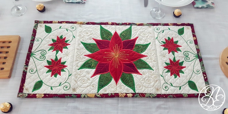 Op de afbeelding: Een witte gestikte tafelrunner met een rood en groen geborduurd kerstster-ontwerp. De runner heeft een bordeauxrode rand met een gouden afwerking.