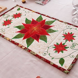 Op de afbeelding: Een witte gestikte tafelrunner met een rood en groen geborduurd kerstster-ontwerp. De runner heeft een rode en gouden rand met een bloemmotief.