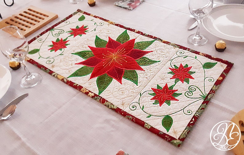 Op de afbeelding: Een witte gestikte tafelrunner met een rood en groen geborduurd kerstster-ontwerp. De runner heeft een rode en gouden rand met een bloemmotief.