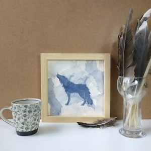 Peut inclure: Une impression encadrée d'un loup hurlant bleu. Un mug à motifs floraux, un vase en verre avec des plumes et une plume unique sont également présents. L'ensemble est disposé sur une surface blanche.