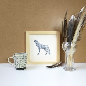 Peut inclure: Une impression encadrée d'un loup hurlant en nuances de gris, une tasse à motifs floraux et un vase en verre rempli de plumes. L'œuvre est exposée sur une surface blanche devant un fond marron, créant une esthétique rustique.