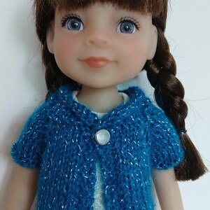 May include: A doll with brown hair styled in two braids, blue eyes, and a light complexion. The doll wears a short-sleeved, knitted blue cardigan with silver metallic threads and a single round white button, layered over a white dress with lace.