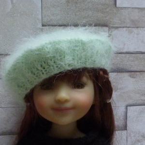 Peut inclure: Un béret en fourrure vert clair avec un motif tricoté délicat. Le béret est porté par une poupée aux longs cheveux bruns et au visage souriant.