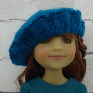 Puede incluir: Una muñeca con una boina de punto azul turquesa y una camisa de manga larga azul turquesa con un gráfico de unicornio.