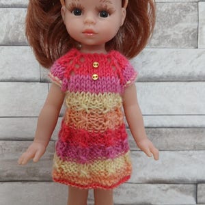 May include: A doll with auburn hair and a peach-colored headband wearing a hand-knitted dress featuring horizontal stripes of pink, yellow, and orange yarn. The dress has a textured stitch pattern, short sleeves, and two small gold buttons at the neckline.
