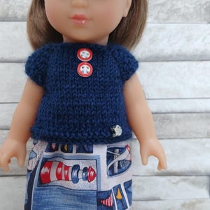 May include: A doll with brown hair and a red headband wearing a navy blue knitted short-sleeved top with two red buttons and a skirt featuring a nautical print of lighthouses and blue patterns, paired with shiny red shoes.