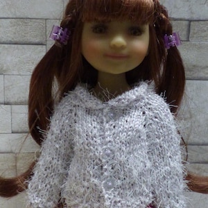 Peut inclure: Une poupée aux cheveux roux coiffée en couettes, retenues par des barrettes violettes. La poupée porte un pull gris clair et duveteux avec des boutons nacrés et un pantalon rose. La poupée a le teint clair et sourit.