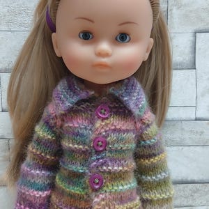 Puede incluir: Una muñeca con cabello rubio y ojos azules que lleva un cárdigan de punto multicolor con botones morados. La muñeca también lleva pantalones morados claros y un lazo morado. El cárdigan presenta tonos de morado, verde, amarillo y marrón.