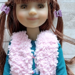 Peut inclure: Poupée aux cheveux bruns tressés, ornés de barrettes violettes. Elle porte un haut à manches longues turquoise, un pantalon en jean et un gilet rose duveteux. Le haut présente un motif de licorne.