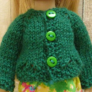 May include: A green knitted cardigan with three green buttons. The cardigan is made for a doll.