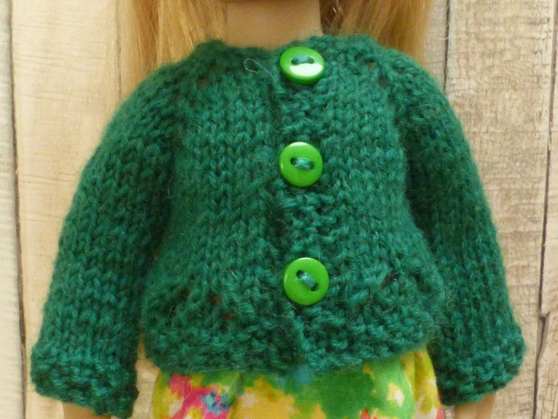 May include: A green knitted cardigan with three green buttons. The cardigan is made for a doll.