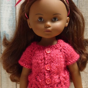 May include: A doll with brown hair wearing a pink knitted cardigan with buttons. The doll has brown eyes and a light brown complexion.