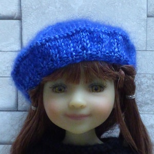 Peut inclure: Une poupée aux longs cheveux bruns portant un béret bleu tricoté. La poupée a un teint clair et porte un pull noir.