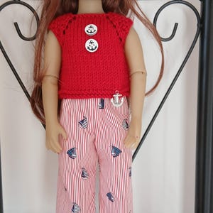 Könnte beinhalten: Eine Puppe mit langem rotbraunem Haar trägt ein rotes, ärmelloses Strickoberteil mit zwei weißen Knöpfen mit Anker-Motiv sowie eine weiße Hose mit feinen roten vertikalen Streifen und einem Muster aus kleinen blau-weißen Segelbooten.