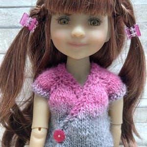 Peut inclure: Une poupée aux cheveux bruns coiffés en couettes, attachées avec des barrettes roses. La poupée porte un haut tricoté rose et gris avec un bouton rouge et un jean bleu. La poupée a le teint clair et regarde le spectateur.