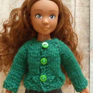 May include: A doll with long brown curly hair wearing a green knitted cardigan with green buttons. The doll is wearing dark blue jeans.